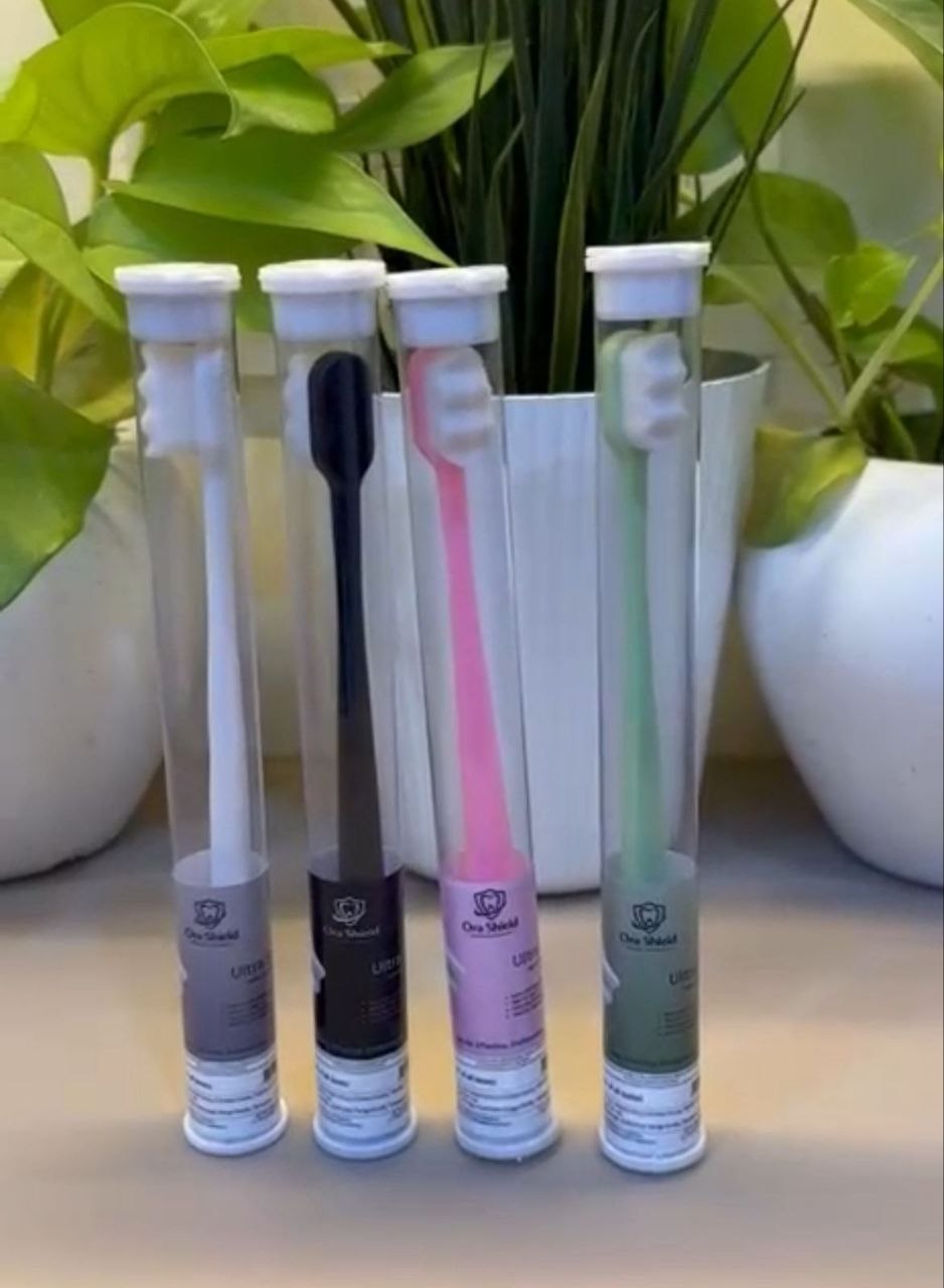 Ora Shield Toothbrushes in 4 colours — white, black, pink, green — in transparent tube packaging beside a plant